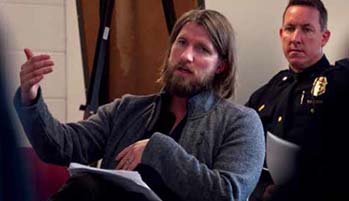 Brad Ray sits in a classroom with a police man behind him.